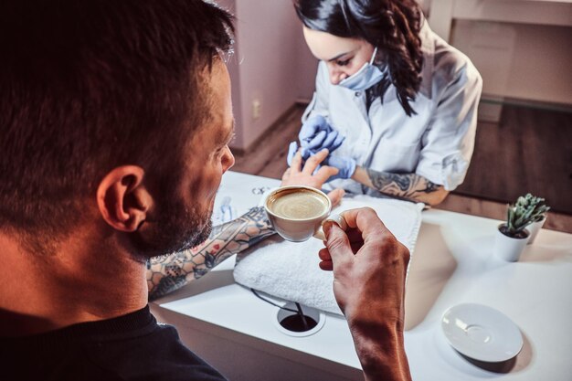 Mężczyzna otrzymujący manicure w gabinecie kosmetycznym, relaksujący się i pijący kawę, podczas gdy mistrz kosmetyczny wykonuje zabieg manicure