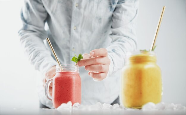 Mężczyzna ozdabia smoothie w rustykalnym słoiku listkami mięty. Dwa słoiki ze zdrowymi, różnymi zimnymi koktajlami owocowymi w środku: mango