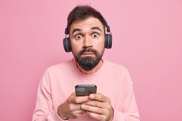 mężczyzna patrzy wyłupiastymi oczami na aparat używa telefonu komórkowego słucha ścieżki dźwiękowej przez bezprzewodowe słuchawki, ubrany w zwykłe ubranie
