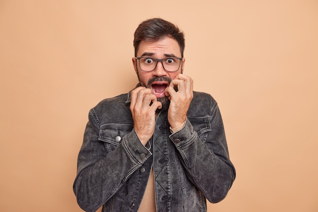 Bezpłatne zdjęcie mężczyzna patrzy z paniką drży ze strachu boi się czegoś trzyma ręce na twarzy trzyma opuszczoną szczękę nosi okulary i dżinsową kurtkę na beżowym studio