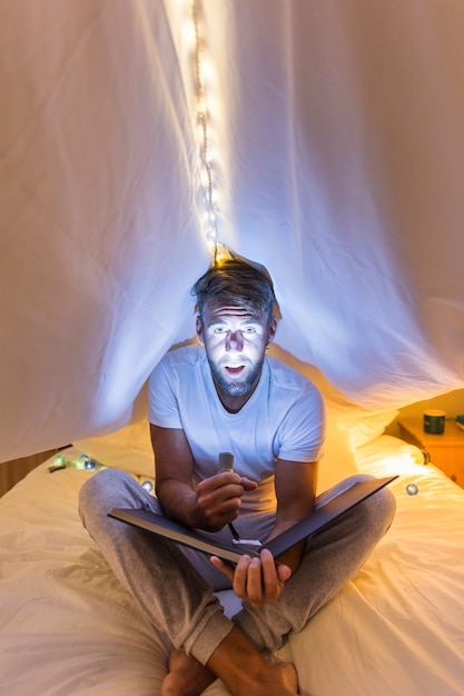 Bezpłatne zdjęcie mężczyzna podkreśla swoją twarz z latarką, siedząc pod zasłoną na łóżku trzymając album
