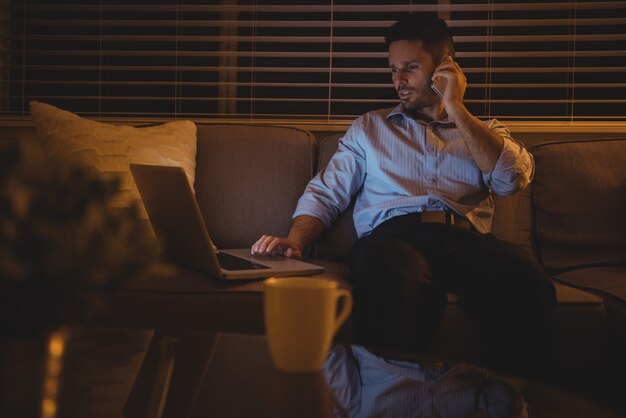 Mężczyzna rozmawia przez telefon komórkowy podczas korzystania z laptopa w salonie