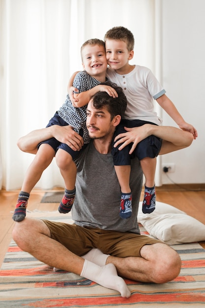 Mężczyzna siedzi na dywanie, niosąc dwóch synów na ramieniu