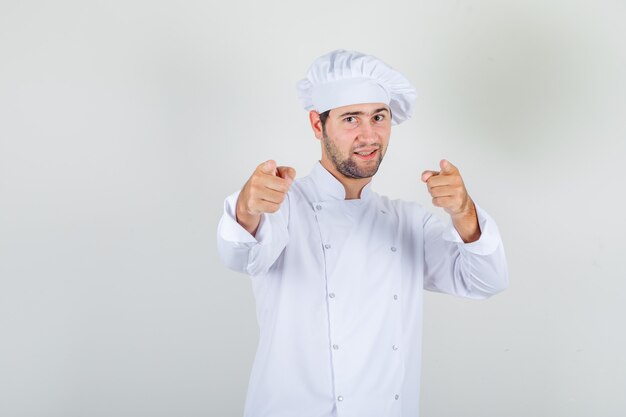 Mężczyzna szefa kuchni wskazując palcami na aparat w białym mundurze i wyglądający wesoło.