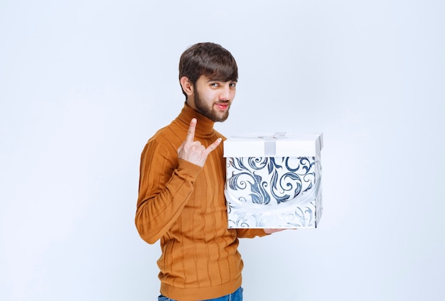 Mężczyzna trzyma białe pudełko z niebieskimi wzorami i pokazuje znak satysfakcji.