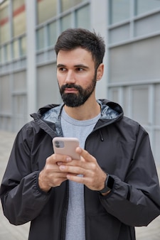 Mężczyzna ubrany w czarną kurtkę używa telefonu komórkowego do czatowania online pobieranie aplikacji do treningu sportowego pozuje na zewnątrz na niewyraźne. nowoczesny styl życia
