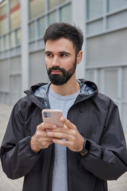 mężczyzna ubrany w czarną kurtkę używa telefonu komórkowego do czatowania online pobieranie aplikacji do treningu sportowego pozuje na zewnątrz na niewyraźne. Nowoczesny styl życia