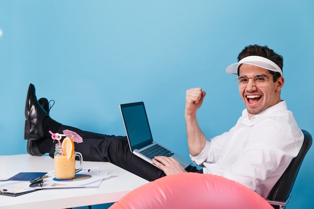 Bezpłatne zdjęcie mężczyzna w czapce i ubraniach biurowych śmieje się podczas pracy i picia koktajlu na niebieskiej przestrzeni. facet trzyma laptopa.