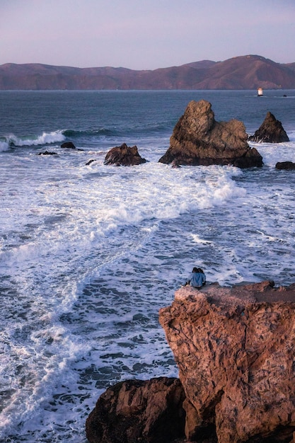 Mężczyzna w czarnej koszuli siedzi na brązowej formacji skalnej w pobliżu morza w ciągu dnia