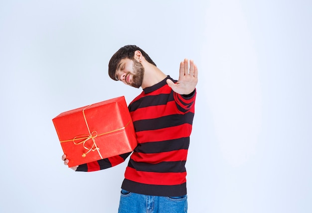 Mężczyzna w czerwonej koszuli w paski, trzymający czerwone pudełko upominkowe, mocno je przytulający i nie chcący się z nikim dzielić.