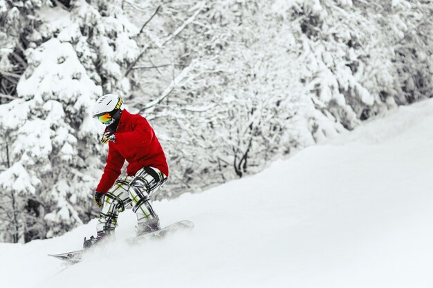 Mężczyzna w czerwonej kurtce narciarskiej i białym hełmie schodzi po zaśnieżonym wzgórzu w lesie