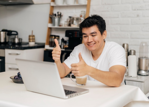 Mężczyzna w domu podczas pandemii pokazujący kciuki do góry podczas rozmowy wideo na laptopie