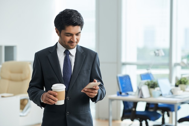 Mężczyzna w garniturze, stojąc w biurze, trzymając kawę na wynos i przy użyciu smartfona
