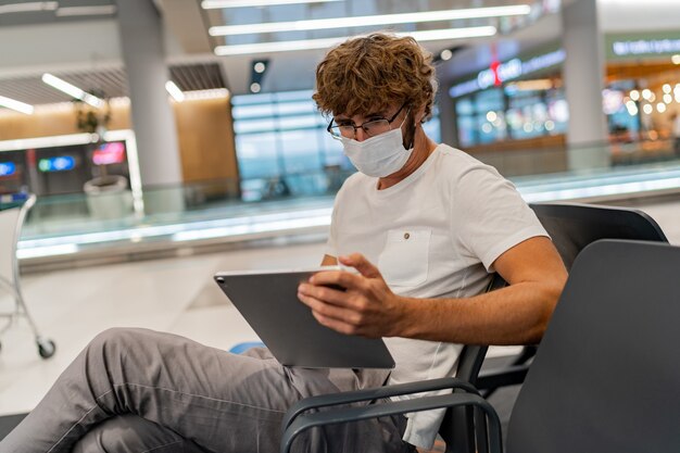 Mężczyzna w masce respiratora czeka na następny samolot na lotnisku i używa tabletu.