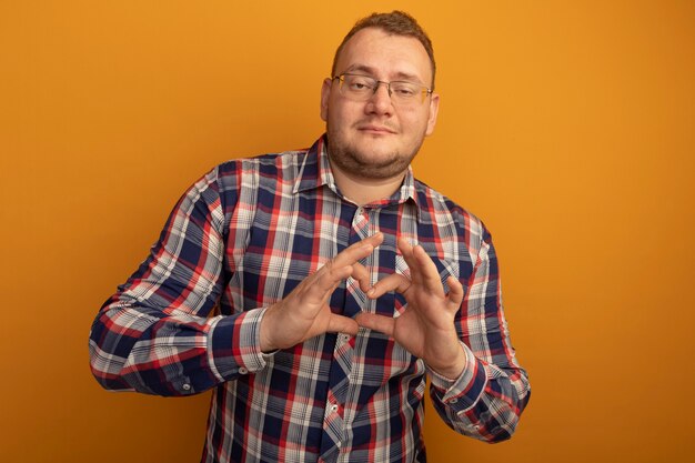 Mężczyzna w okularach i koszuli w kratkę robi gest serca palcami uśmiechając się pewnie stojąc nad pomarańczową ścianą