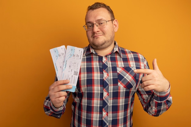 Mężczyzna w okularach i koszuli w kratkę trzymający bilety lotnicze wskazujący na nich palcem wskazującym, uśmiechnięty pewnie, stojący nad pomarańczową ścianą