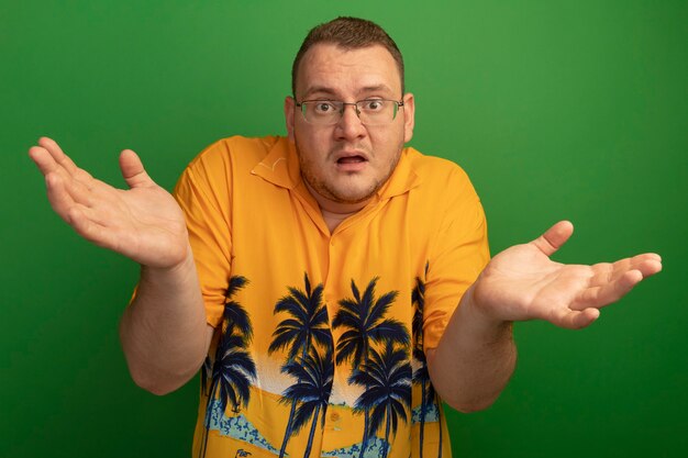 Bezpłatne zdjęcie mężczyzna w okularach i pomarańczowej koszuli mylić z podniesionymi rękami stojąc nad zieloną ścianą