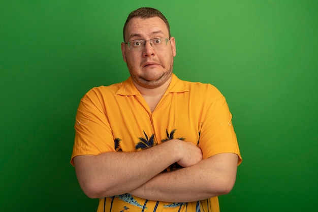 Bezpłatne zdjęcie mężczyzna w okularach i pomarańczowej koszuli mylić ze skrzyżowanymi rękami stojąc nad zieloną ścianą
