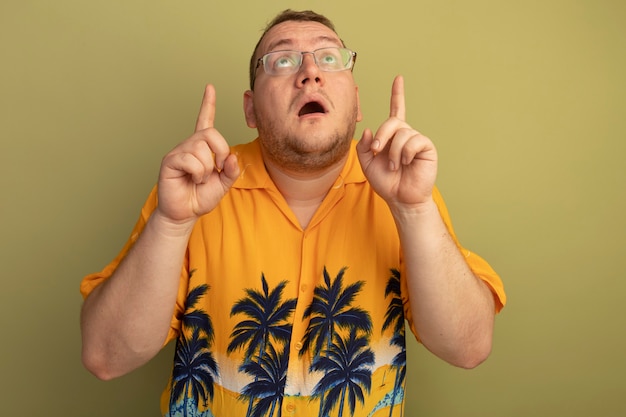 Bezpłatne zdjęcie mężczyzna w okularach w pomarańczowej koszuli patrząc w górę zaskoczony i zmartwiony wskazując palcami wskazującymi w górę, stojąc nad zieloną ścianą