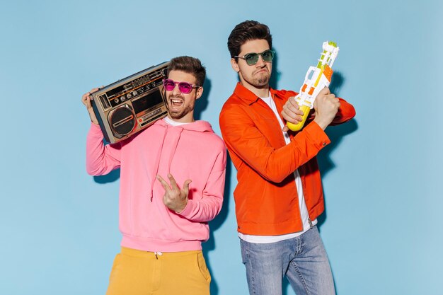 Mężczyzna w różowej bluzie i okularach przeciwsłonecznych trzyma gramofon Facet w pomarańczowej kurtce i dżinsach bawi się pistoletem na wodę Przyjaciele bawią się na niebieskim tle