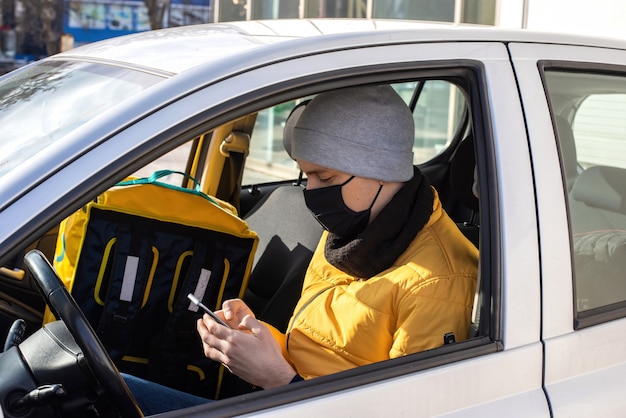 Mężczyzna w samochodzie w czarnej masce medycznej siedzi przy telefonie z plecakiem na siedzeniu