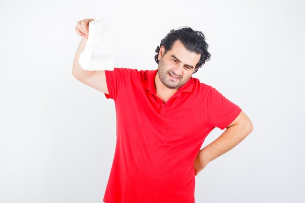 Mężczyzna w średnim wieku podnosząc serwetkę, trzymając rękę na biodrze w czerwonej koszulce i patrząc zamyślony, widok z przodu.