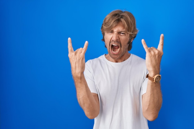 Bezpłatne zdjęcie mężczyzna w średnim wieku stojący na niebieskim tle krzyczący z szalonym wyrazem twarzy, wykonujący symbol rocka z rękami w górze, gwiazdą muzyki, koncepcją ciężkiej muzyki