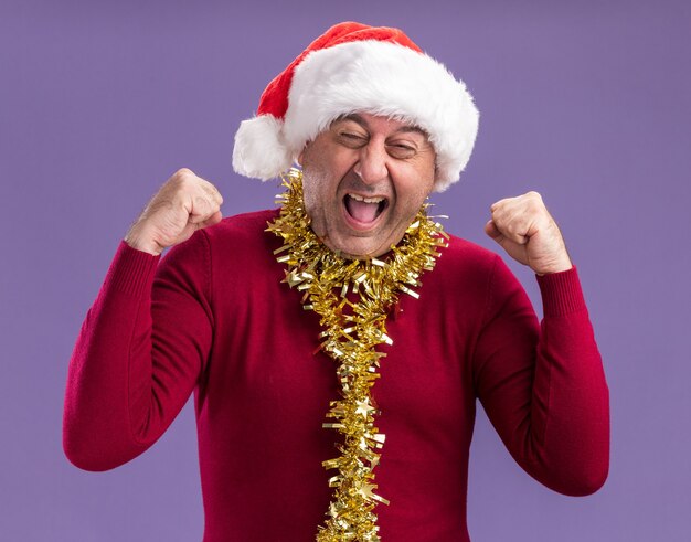 Mężczyzna w średnim wieku w świątecznej czapce mikołaja z blichtrem na szyi szczęśliwy i podekscytowany zaciskając pięści krzycząc stojąc na fioletowym tle