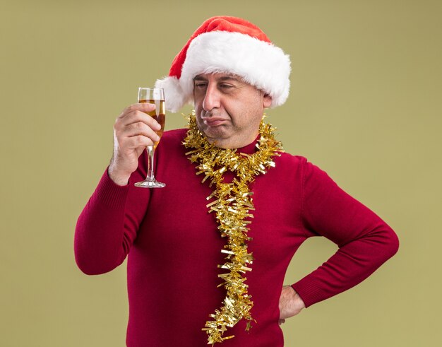 Mężczyzna w średnim wieku w świątecznej czapce mikołaja ze świecidełkiem na szyi trzyma kieliszek szampana, patrząc na niego ze sceptycznym wyrazem twarzy, stojąc na zielonym tle