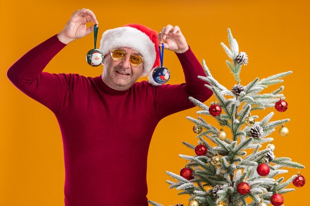 mężczyzna w średnim wieku w świątecznym czapce mikołaja w ciemnoczerwonym golfie i żółtych okularach trzyma bombki uśmiechnięte wesoło stojąc obok choinki na pomarańczowym tle