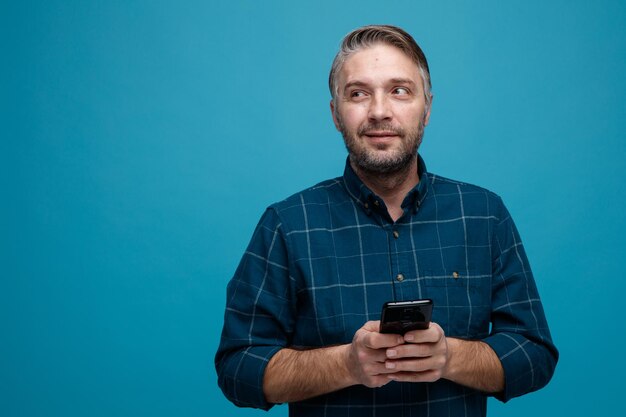 Mężczyzna w średnim wieku z siwymi włosami w ciemnej koszuli trzymający smartfona patrzący w bok z zamyśloną miną myślący uśmiechnięty stojący na niebieskim tle