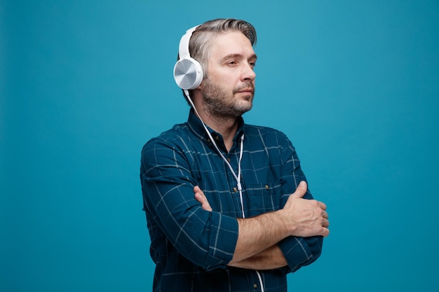 Mężczyzna w średnim wieku z siwymi włosami w ciemnej koszuli ze słuchawkami, patrzący na bok z poważną twarzą z rękami skrzyżowanymi na piersi, stojący na niebieskim tle