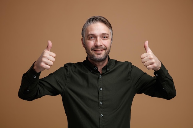 Mężczyzna W średnim Wieku Z Siwymi Włosami W Ciemnozielonej Koszuli Patrzący Na Kamerę Szczęśliwy I Pozytywny Uśmiechnięty Pokazujący Kciuki Do Góry Stojący Na Brązowym Tle Premium Zdjęcia