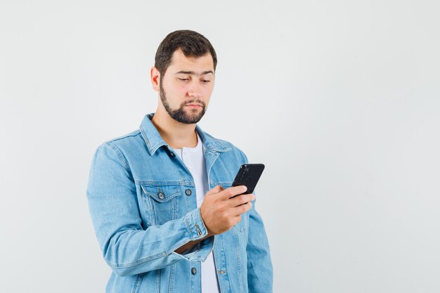 Mężczyzna w stylu retro czyta coś na telefon komórkowy w kurtce, t-shirt i patrząc skoncentrowany, widok z przodu.