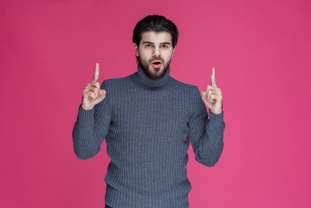 Mężczyzna w szarym swetrze wskazujący coś lub przedstawiający kogoś palcem wskazującym.