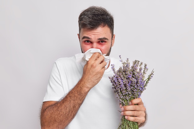 Bezpłatne zdjęcie mężczyzna wydmuchuje nos w chusteczce ma kichanie i katar uczulenie na lawendowoczerwone opuchnięcie oczu cierpi na nieprzyjemne objawy na białej ścianie. pojęcie choroby