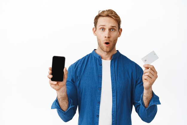 Mężczyzna wygląda na zaskoczonego, pokazuje ekran telefonu komórkowego i kartę kredytową, mówiąc o funkcji banku, ofercie zakupów online, stojąc zdumiony na białej ścianie