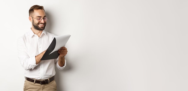 Bezpłatne zdjęcie mężczyzna wyglądający na zadowolonego podczas czytania dokumentów, trzymający schowek i uśmiechający się, stojąc nad białymi plecami