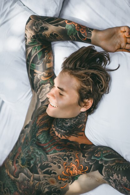 Mężczyzna z kolorowymi tatuażami pozuje na białym prześcieradle