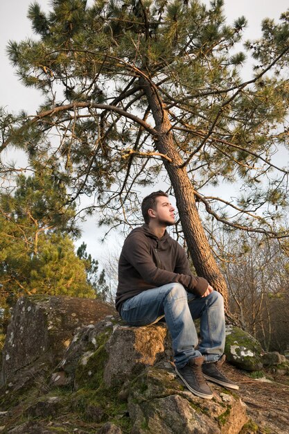 Mężczyzna z plecakiem siedzący na skałach w lesie i patrzący na horyzont w ponury dzień