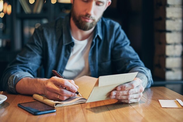 Mężczyzna z ręką pióra piszący na notatniku na drewnianym stole. Mężczyzna pracujący w kawiarni.