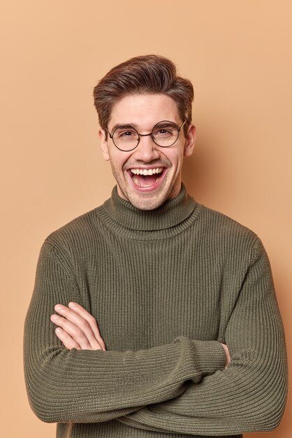 mężczyzna z wyrazem twarzy zadowolenia trzyma założone ręce ma pozytywną rozmowę z przyjacielem przedyskutuj coś śmiesznego nosi ciepły sweter pozuje na beżowym.