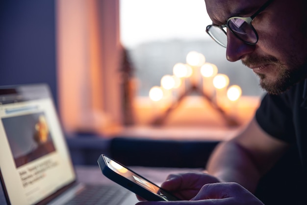 Bezpłatne zdjęcie mężczyzna ze smartfonem siedzi przed laptopem późno w nocy