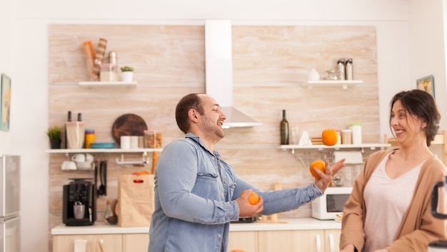 Mężczyzna żongluje pomarańczami dla kobiety, przygotowując smaczne i pożywne smoothie. Zdrowy beztroski i wesoły tryb życia, dieta i przygotowanie śniadania w przytulny słoneczny poranek