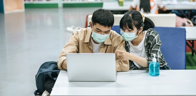 Mężczyźni i kobiety noszą maski i używają laptopa do wyszukiwania książek w bibliotece.