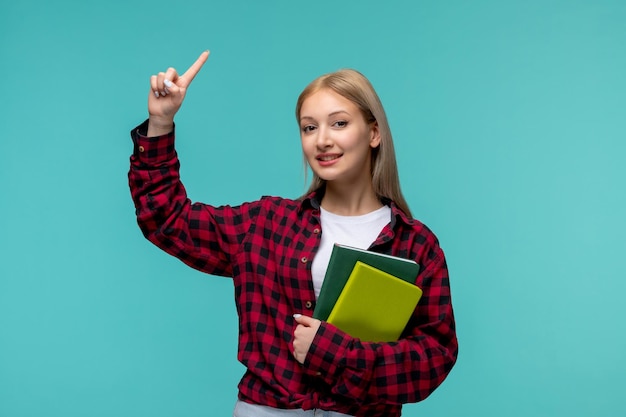 Bezpłatne zdjęcie międzynarodowy dzień studencki blondynka ładna dziewczyna w czerwonej koszuli w kratę szczęśliwie wskazując palcem