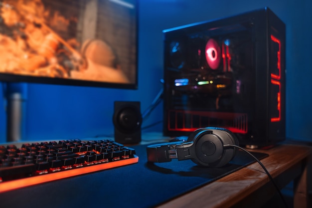 Miejsce pracy dla graczy komputerowych z nową klawiaturą do gier, myszą, słuchawkami, nowoczesnym komputerem pc z rozmytym niebieskim i czerwonym światłem neonowym.