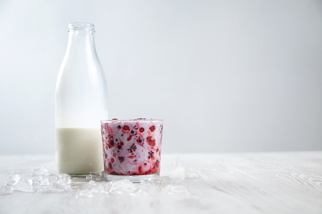 Mieszane mrożone jagody w szkle rox zmieszane z organicznym świeżym mlekiem w pobliżu rocznika butelki na białym tle otoczone stopionymi kostkami lodu