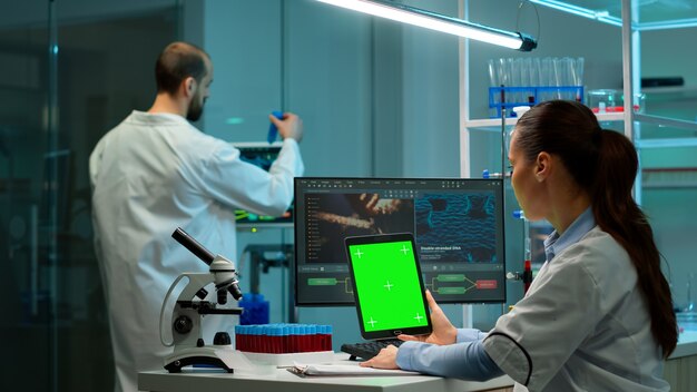 Mikrobiolog pracujący na notatniku z zielonym wyświetlaczem chroma key w nowocześnie wyposażonym laboratorium. Zespół biotechnologów opracowujących leki przy użyciu tabletu z makietą ekranu.