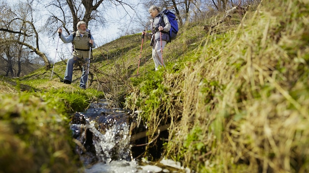 Miłość i jedność. Starsza rodzina para mężczyzna i kobieta w stroju turystycznym spaceru na zielonym trawniku w pobliżu drzew i potoku w słoneczny dzień